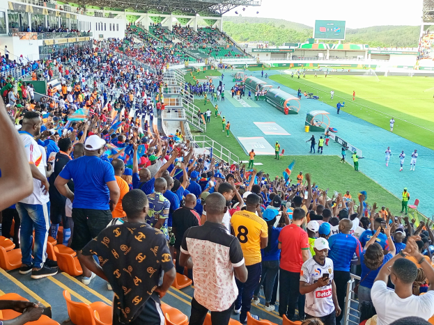 Eine große Menschenmenge versammelt sich vor einem eingezäunten Fußballfeld, wobei einige auf Stühlen sitzen und andere stehen, mit Blick auf Hügel und einen klaren blauen Himmel während der African Cup of Nations 2019.