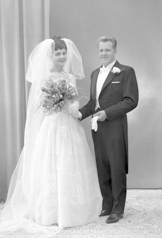 Ein Schwarz-Weiß-Foto eines strahlenden Paares an ihrem Hochzeitstag, wobei die Frau in einem weißen Kleid mit einem Bukett in der Hand und einer Vorhangwand im Hintergrund zu sehen ist.