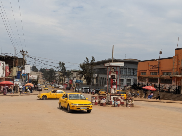Ein gelbes Taxi fährt eine belebte Stadtstraße mit Gebäuden an den Seiten hinunter, während Fußgänger mit Schirmen auf dem Gehweg unter wolkenverhangenem Himmel gehen.