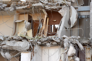 Ein teilweise abgerissener Bau mit zerbrochenen Wänden und verstreuten Trümmern, darunter Holzstücke, in einem verwahrlosten Zustand.