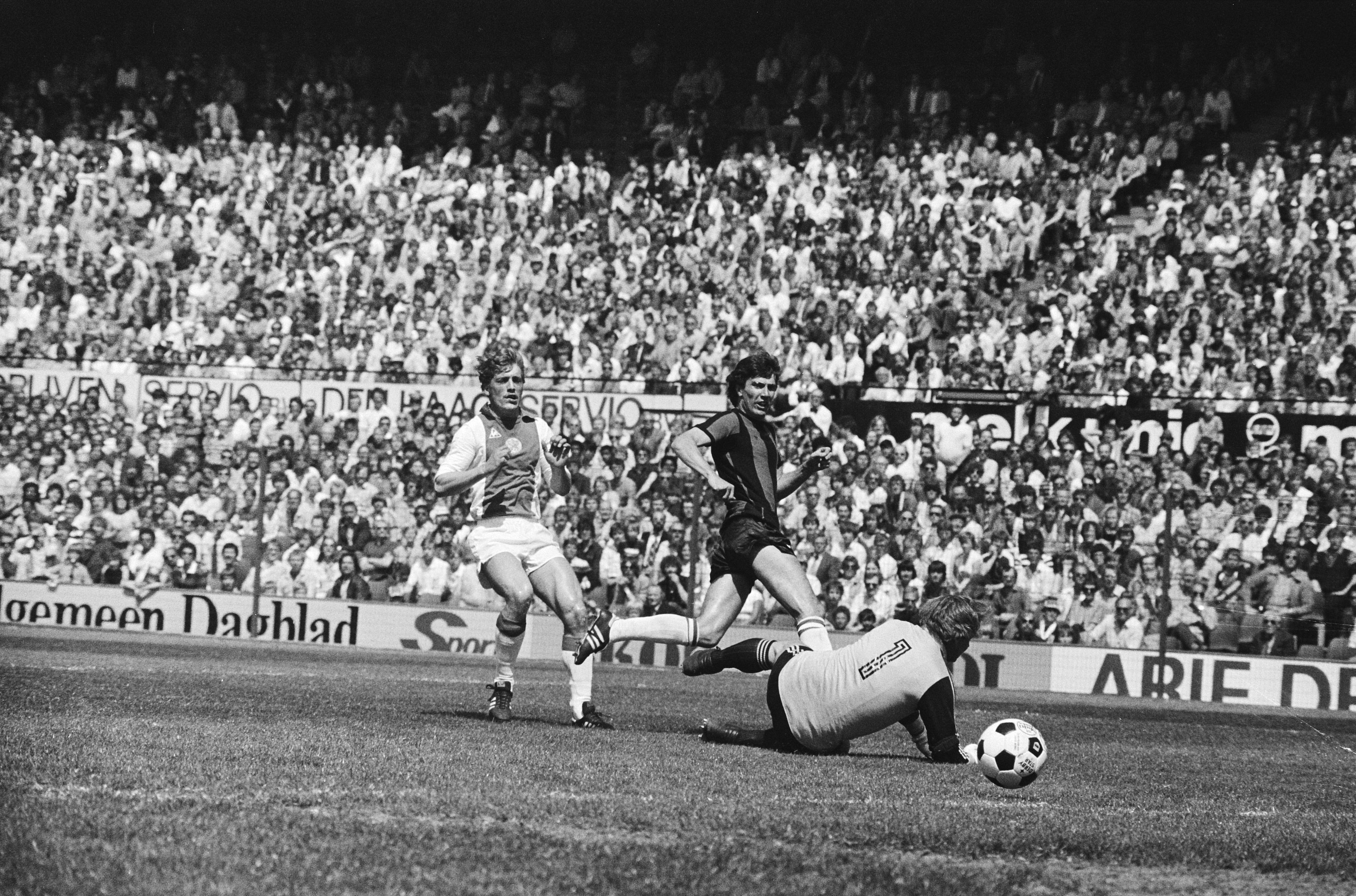 Schwarzes und weißes Foto von Männern, die auf einem Feld Fußball spielen, mit Zuschauern im Hintergrund und einer "Bundesliga 1978-79 Ajax 1-1 Hannover 96"-Plakate.