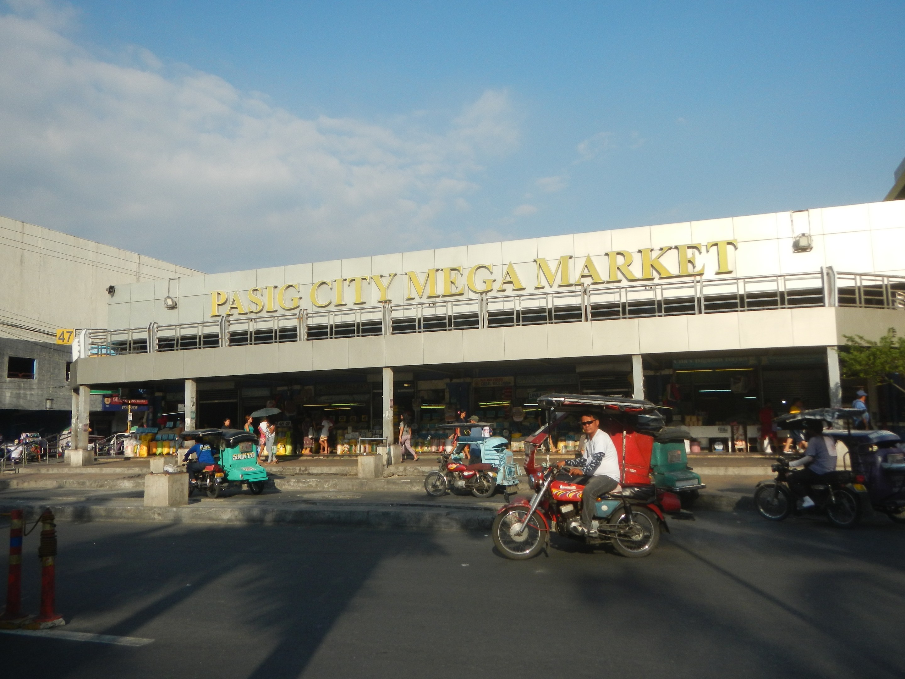 Eine belebte Pasig City Mega Market Szenerie mit Fahrzeugen auf der Straße und Fußgängern, ein Gebäude mit Text im Hintergrund und ein bewölkter Himmel.