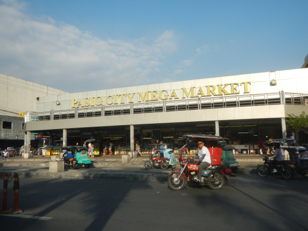 Eine belebte Pasig City Mega Market Szenerie mit Fahrzeugen auf der Straße und Fußgängern, ein Gebäude mit Text im Hintergrund und ein bewölkter Himmel.