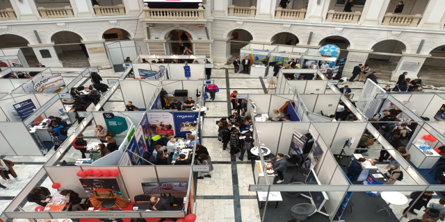 Eine große Gruppe von Menschen versammelte sich in einem Raum voller Stände, Banner, Tische, Stühle und Ballons, mit einem Gebäude im Hintergrund mit Bögen, Säulen, Geländern und einem Bildschirm.