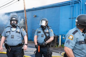 Drei Polizisten in Einsatzausrüstung, darunter Helme und Gasmasken, gehen eine Straße mit einer blauen Wand, Rohren und einem Strommast im Hintergrund entlang.