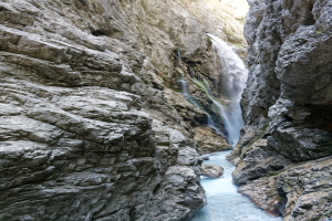 Kleiner Wasserfall, der über schroffe Felsen in einem steinigen Tal hinabstürzt, umgeben von saftig grünen Hügeln unter strahlendem Sonnenschein.