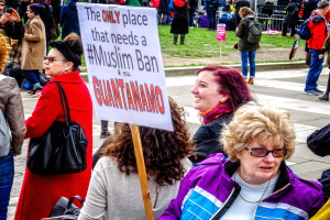 Eine Gruppe von Menschen steht auf dem Boden, einige halten Taschen, eine Frau hält ein Schild mit der Aufschrift "Der einzige Ort, der ein muslimisches Verbot benötigt, ist Guantanamo", vor einem Metallzaun, Gras, einem Schild, Pfosten, einem Gebäude mit Fenstern und Bäumen.