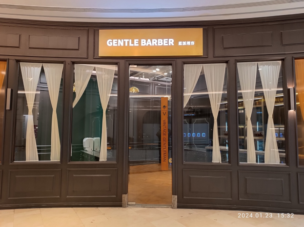 Ein Friseursalon mit Glastüren und Vorhängen, einer Wandtafel, Deckenleuchten und einem Wasserzeichen unten, Located in einem Einkaufszentrum.