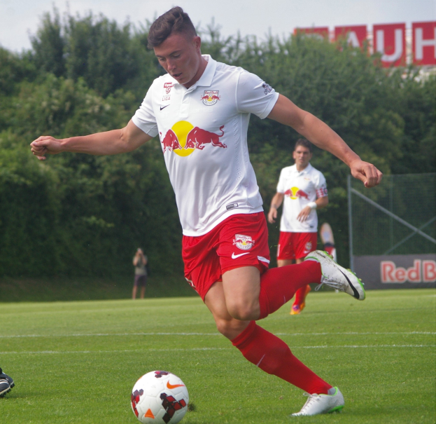 Ein Mann in einem Fußballtrikot kickt einen Ball auf einem grünen Feld mit Bäumen und einem klaren blauen Himmel im Hintergrund, mit einem Schild mit der Aufschrift "RB Leipzig" dahinter.